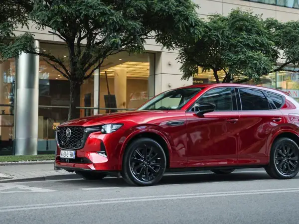 Lśniąca, czerwona Mazda CX-60 3.3 diesel na tle nowoczesnego budynku i drzew.