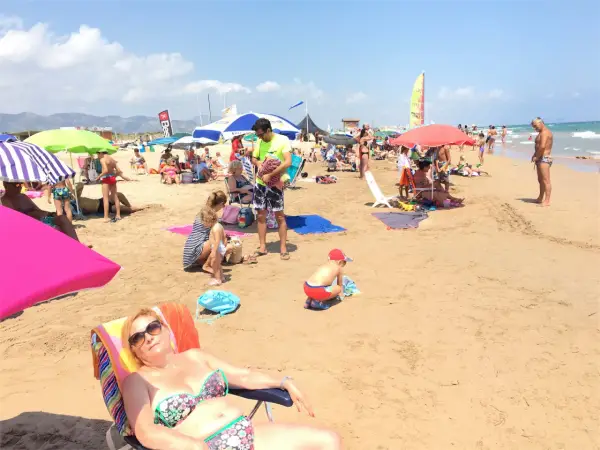 Día soleado en la playa de l'Ahuir, con gente disfrutando bajo sombrillas y el mar al fondo.