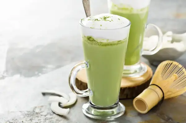 Dwa zielone napoje matcha w szklankach z pianką, obok bambusowa miotełka i wiórki kokosowe. Tak smakuje matcha.