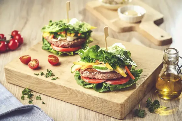 Dwa soczyste burgery w sałacie, z pomidorami, ogórkiem i papryką, na drewnianej desce.