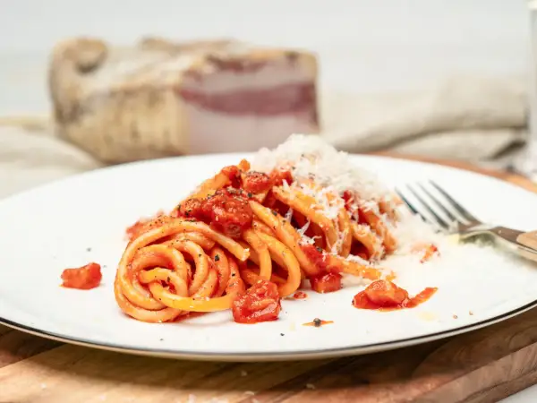 Ein Teller Pasta all'Amatriciana, garniert mit geriebenem Parmesan. Im Hintergrund ein Stück Guanciale, die Zutat für das authentische Amatriciana Rezept.