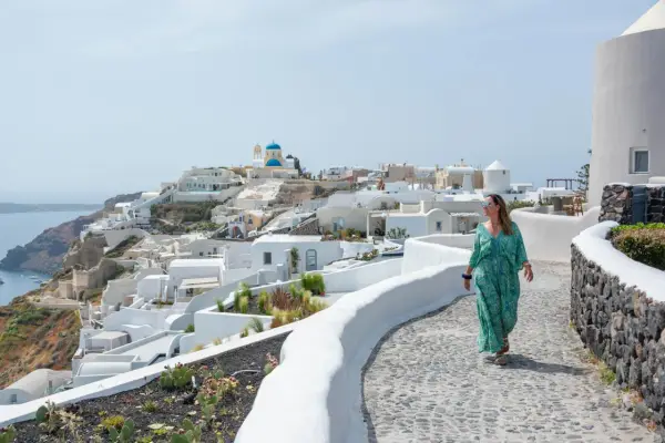 Kobieta w sukience spaceruje po Santorini, idealnym miejscu na wakacje w Grecji. Białe domy i błękitna kopuła kościoła tworzą malowniczy krajobraz.