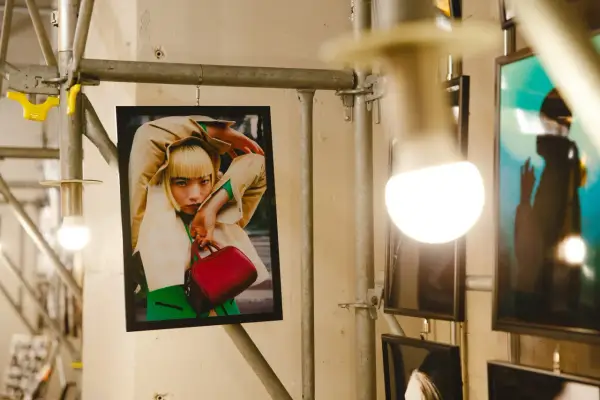 Plakat z kobietą w beżowym płaszczu i zielonym swetrze, trzymającą czerwoną torebkę. Stylizacja przypomina modę z **Pezet Tokyo**.