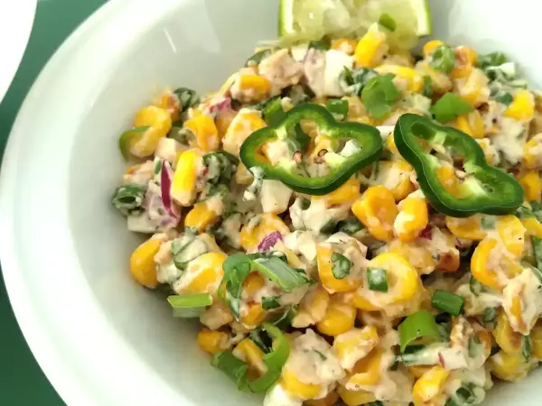 Deliciosa ensalada de maiz cremosa con jalapeños, cebolla morada y cebollín, servida en un tazón blanco.