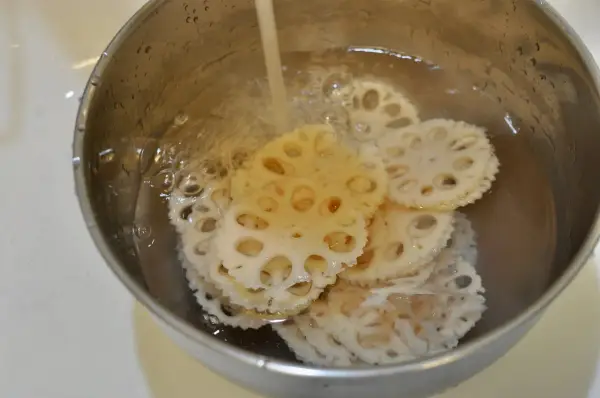 Slices of pickled lotus root are being rinsed in a metal bowl under running water, ready for a delicious meal.