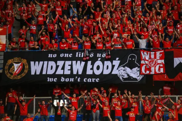 Tłum kibiców Widzewa w czerwonych koszulkach skanduje przyspiewki na Widzew, machając rękami. Na banerze widnieje napis "WIDZEW ŁÓDŹ".