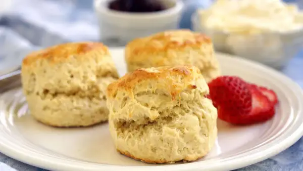 Tre gyllene engelska scones på en vit tallrik med en jordgubbe, sylt och grädde i bakgrunden.