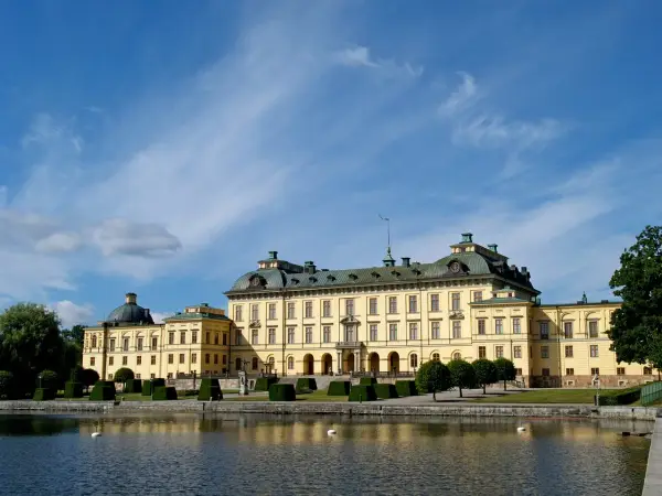 Drottningholm slott, ett gult barockpalats med gröna tak, speglar sig i vattnet under en blå himmel med tunna moln.
