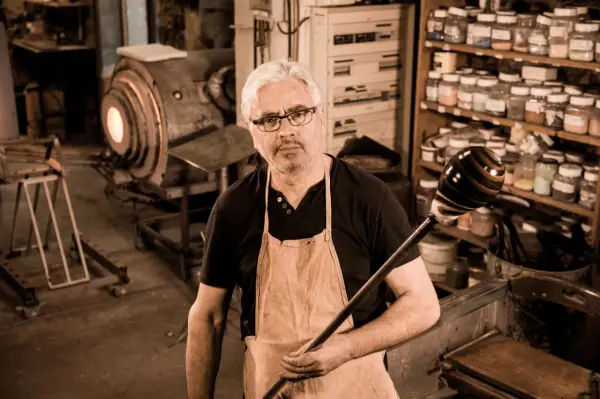Un artisan verrier expérimenté, tablier en cuir, tient sa canne à souffler devant son four incandescent.
