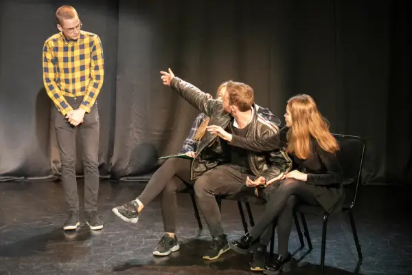 Młodzi aktorzy ćwiczą scenę. Jeden z nich wskazuje coś na widowni, jakby to był teatr na bruku.