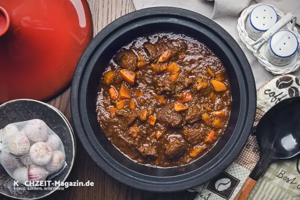 Herzhaftes Rehhals Rezept in einem Schmortopf, mit Karottenstücken und Knoblauch daneben.