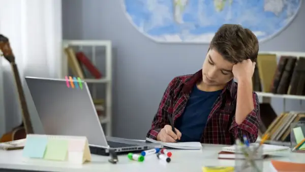 Ein Junge sitzt frustriert am Schreibtisch und macht seine hausaufgaben. Ein Laptop und bunte Stifte liegen bereit.