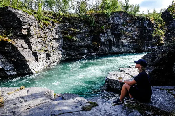 En kvinna sitter vid en forsande turkos flod i en klippig ravin. Vacker natur, kanske en vy över var ligger Abisko.