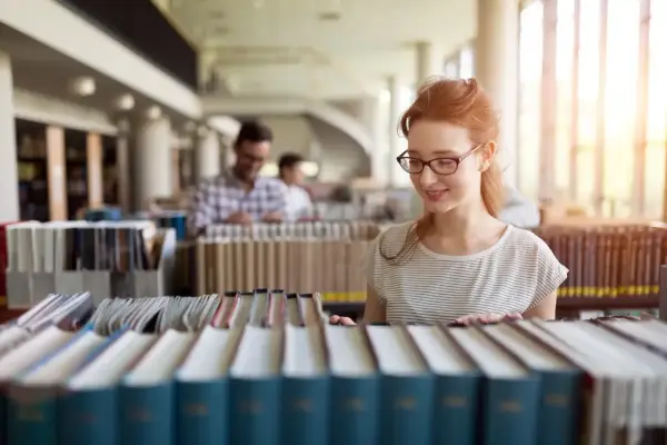 Studentka z rudymi włosami w okularach przegląda książki w bibliotece, szukając materiałów do prawo studia.