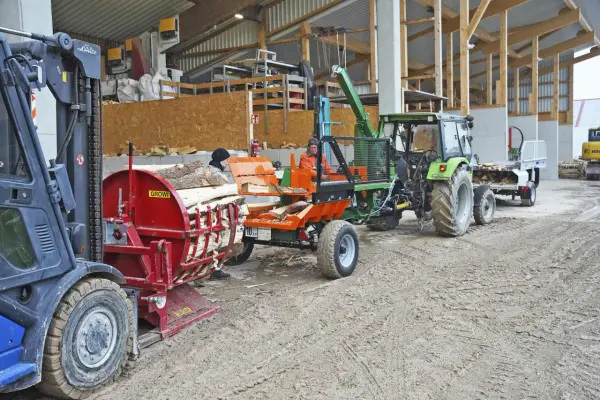 Gabelstapler lädt Holz auf eine hydraulische Kreissäge. Ein Traktor mit Anhänger wartet daneben.