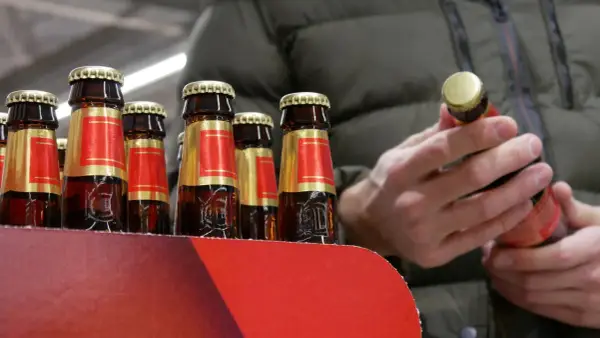 Rząd butelek piwa w czerwono-złotych etykietach. Człowiek w zielonej kurtce trzyma jedną butelkę, zastanawiając się, ile kosztuje jedno piwo.