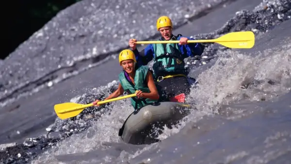 Dwóch uśmiechniętych ludzi w kaskach i kamizelkach pokonuje rwący nurt rzeki na pontonie. To właśnie rafting – emocjonująca przygoda!