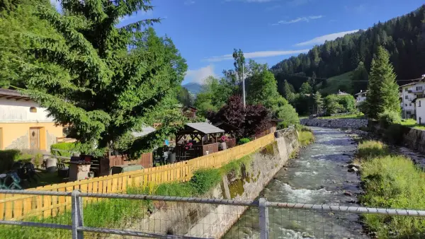 Un ruscello scorre a Santo Stefano di Cadore, fiancheggiato da case, alberi e un'area con gazebo e tavoli all'aperto.