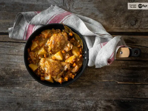 Estofado de pollo con patatas y zanahorias en sartén sobre mesa de madera.
