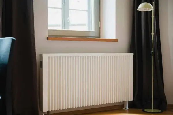 Biały grzejnik pod oknem, obok ciemnej zasłony i lampy stojącej. Dowiedz się, jak ustawić grzejniki przy ogrzewaniu gazowym dla optymalnego komfortu.