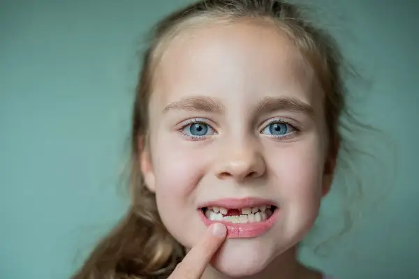 Uśmiechnięta dziewczynka z niebieskimi oczami pokazuje brakujący ząbek. Czy mleczaki mają korzenie? Tak, i zaraz wyrośnie nowy!