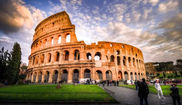 Il Colosseo, un'icona di cosa visitare a Roma, si erge maestoso sotto un cielo drammatico. Turisti passeggiano sul selciato, ammirando l'antica grandezza.