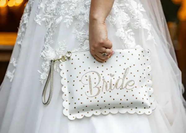 Biała kopertówka w złote kropki z napisem "Bride" to idealny niezbędnik pary młodej. Dopełnia suknię ślubną.