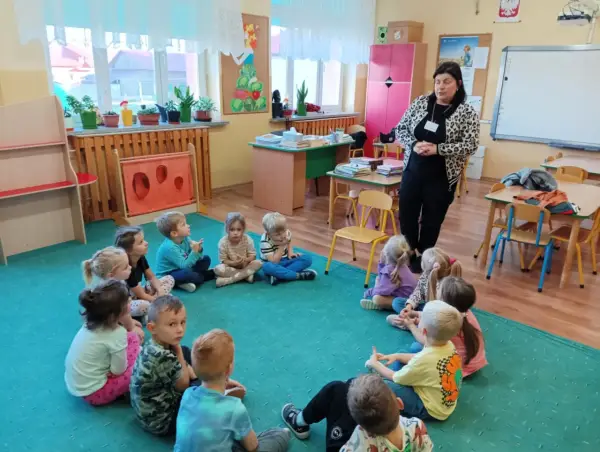 Kobieta w panterkowej kurtce stoi przed grupą dzieci siedzących w kręgu na zielonej wykładzinie.