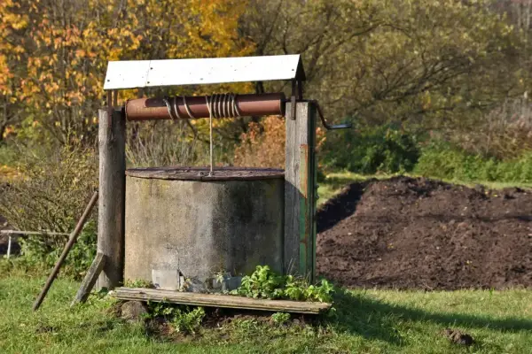 Stary, betonowy krąg studni kręgowej, z drewnianą konstrukcją i metalową korbą, otoczony jesienną zielenią.