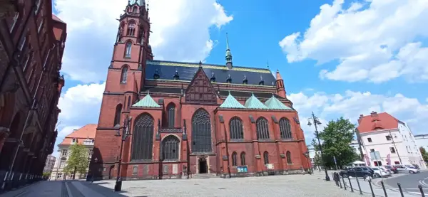Gotycka katedra w mieście nad Kaczawą, z ceglaną fasadą i zielonymi dachami, na tle błękitnego nieba z chmurami.