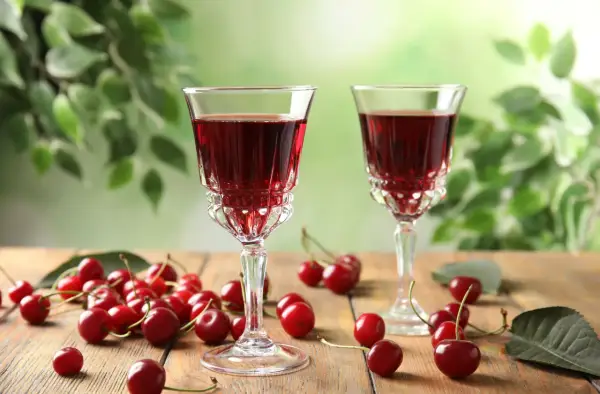 Dwa kieliszki z rubinowym alkoholem z wiśni, otoczone świeżymi owocami i zielonymi liśćmi.