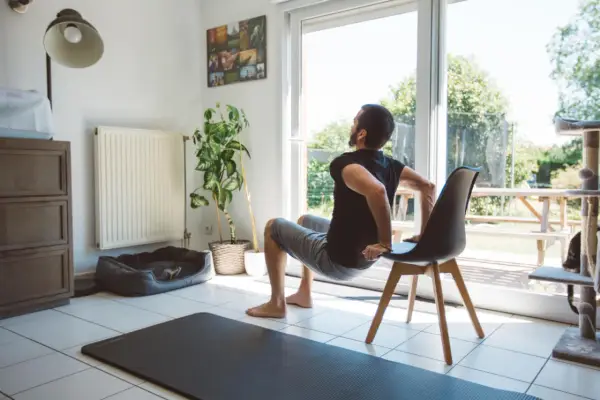 Un homme fait des dips sur une chaise, une idée de quel sport faire à la maison. Un tapis de yoga est au sol.