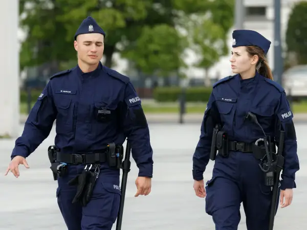 Dwóch policjantów w mundurach idzie, jeden z nich ma na pasie rękawiczki i pałkę.