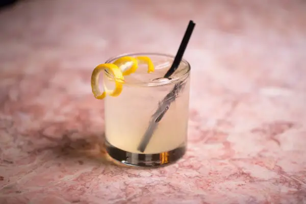 Un rinfrescante Vodka Sour IBA, guarnito con scorza di limone, su un tavolo di marmo rosa.
