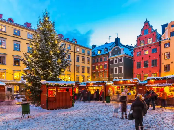 En snötäckt vinterviken julmarknad med ett stort, upplyst träd och färgglada hus. Folk strosar bland bodarna.