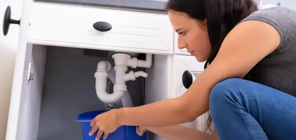 Une femme rattrape une fuite sous un évier dans un seau bleu. Problème de plomberie, que ce soit pour un locataire ou un propriétaire.