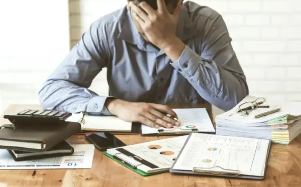 Zestaw dokumentów, kalkulator i telefon leżą na biurku. Mężczyzna z ręką na czole, przytłoczony problemami finansowymi, przegląda pismo windykacyjne.
