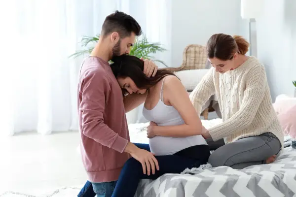 Mężczyzna obejmuje ciężarną kobietę, która opiera głowę na jego ramieniu. Obok siedzi doula, która delikatnie dotyka jej brzucha, oferując wsparcie.