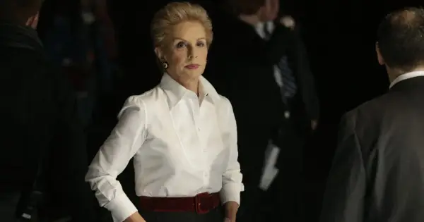 Elegancka Carolina Herrera w białej koszuli i czerwonym pasie, otoczona przez ludzi w ciemnych strojach.
