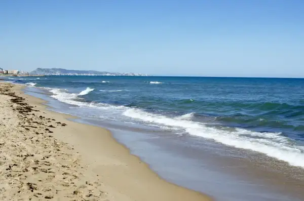 La playa de Xeraco, con arena dorada y el mar azul, se extiende hacia un horizonte con edificios y montañas.