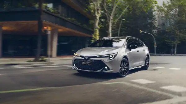 Srebrna Toyota Corolla kombi z czarnymi felgami sunie po miejskiej ulicy, otoczona zielenią i nowoczesną architekturą.
