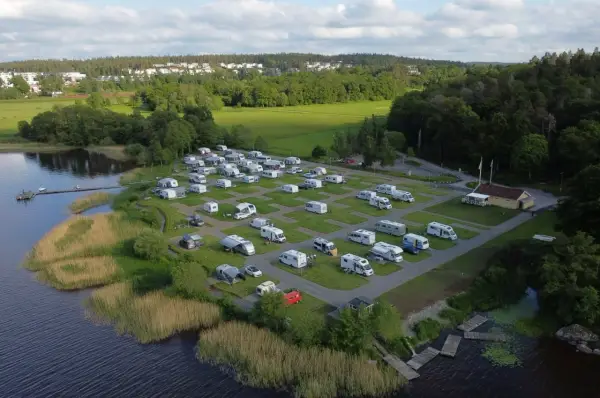 Full campingplats med husbilar och husvagnar vid en sjö, med grönskande träd och bebyggelse i bakgrunden. En idyllisk plats för camping i Alingsås.