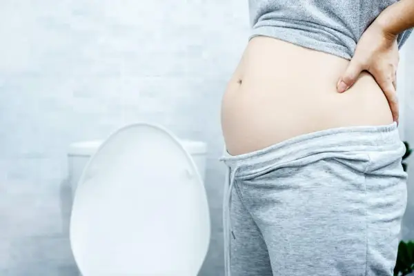 Frau mit Blähbauch hält sich den Bauch. Was hilft gegen Blähbauch? Die Toilette im Hintergrund deutet auf Verdauungsprobleme hin.