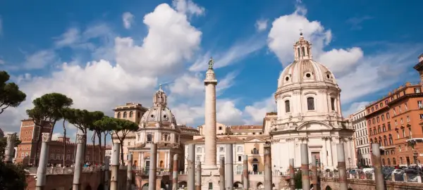 La colonna di Traiano si erge maestosa a Roma, con cupole barocche e pini marittimi sotto un cielo azzurro e nuvoloso.