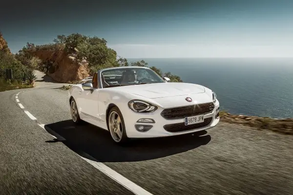 Biały Fiat 124 Spider sunie po krętej drodze nad morzem.