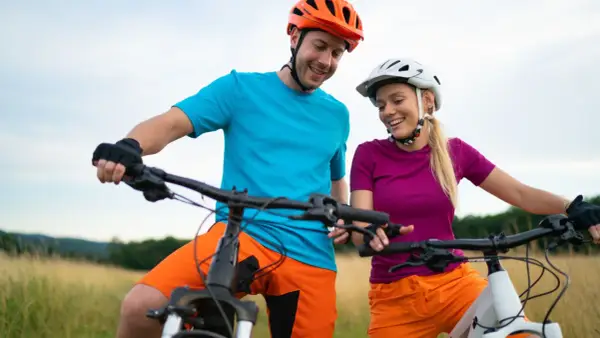 Uśmiechnięta para w kaskach i sportowych strojach, gotowa na przejażdżkę rowerami elektrycznymi. Wygodne akcesoria do roweru elektrycznego zapewniają komfort.