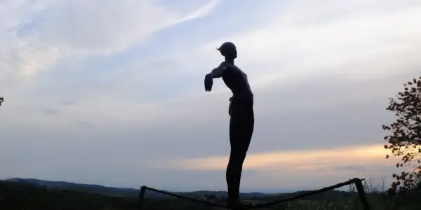 Statua silhouette con braccia aperte, su un crinale collinare, al tramonto. Un punto panoramico lungo le tappe della Via Romea Sanese.
