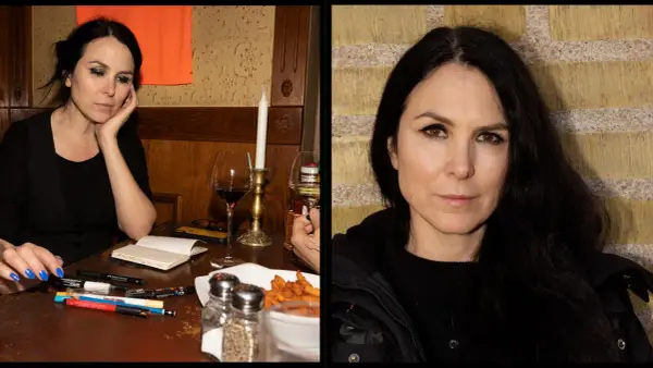 Nina Hemmingsson, med sitt karakteristiska uttryck, sitter vid ett bord med pennor och ett glas rödvin.