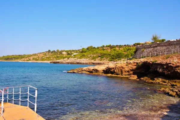 Scogliera rocciosa e mare cristallino a San Gregorio al mare, con vegetazione mediterranea e un muretto in pietra.