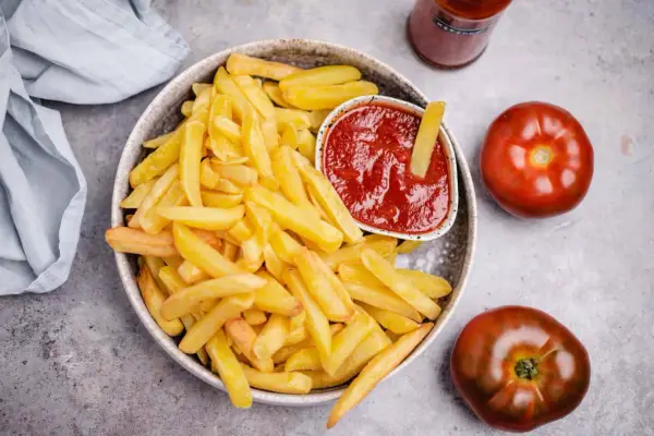 Knusprige Pommes Frites mit einem Schälchen veganem Ketchup. Zwei Tomaten und eine Flasche Ketchup liegen daneben.